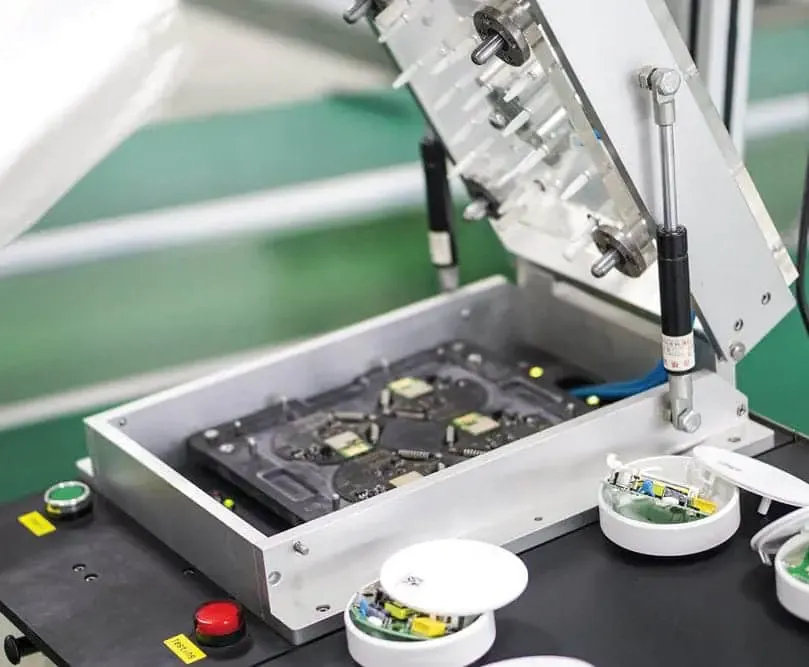 Open electronic test bench holding a circuit board, next to round sensors being assembled