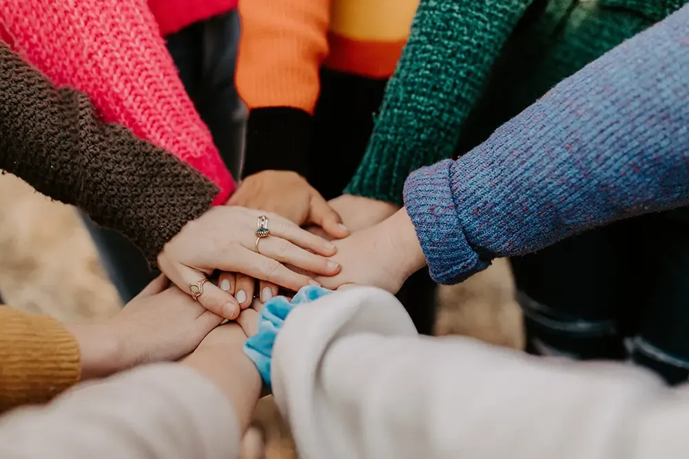Hands of several people in colourful jumpers stacked on top of each other in the middle of a circle