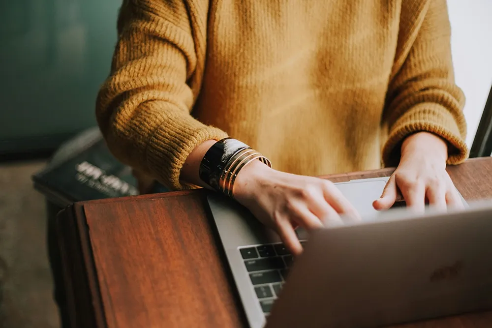 Personne en pull moutarde tapant sur un ordinateur portable posé sur un bureau en bois