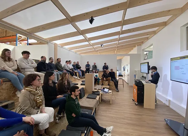 Employees seated on wooden tiered benches in a room with an exposed timber roof, listening to a presentation shown on a screen
