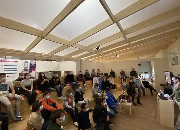 Masked employees seated on wooden tiered benches, listening to a presenter in a room with an exposed timber roof